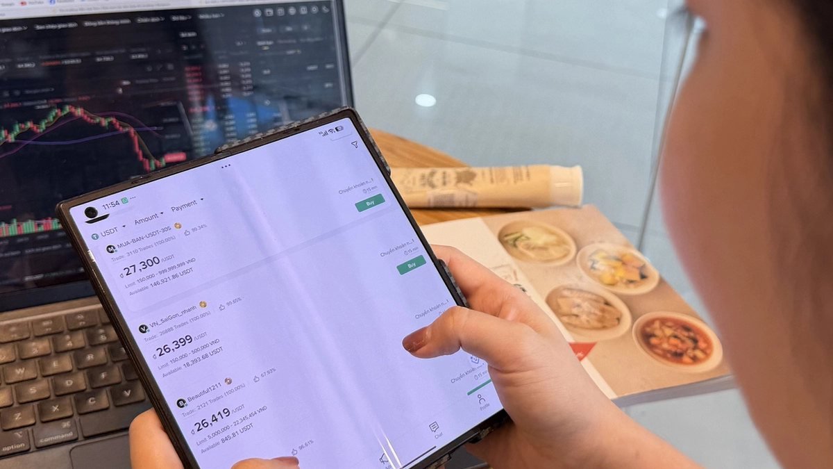 Trader checking open positions on a foldable phone, with a price chart open on the laptop behind, at a café table in Vietnam