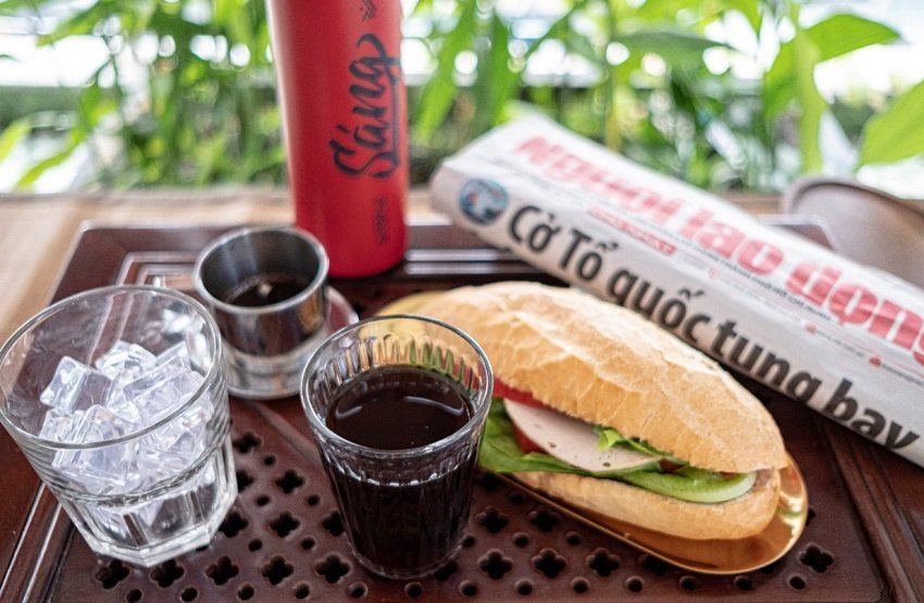 A bowl of pho, a banh mi and an iced Vietnamese coffee on a café table next to a closed laptop